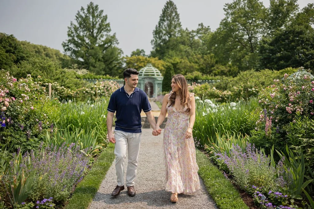 A couple holding hands and walking through a lush garden with blooming flowers on either side.