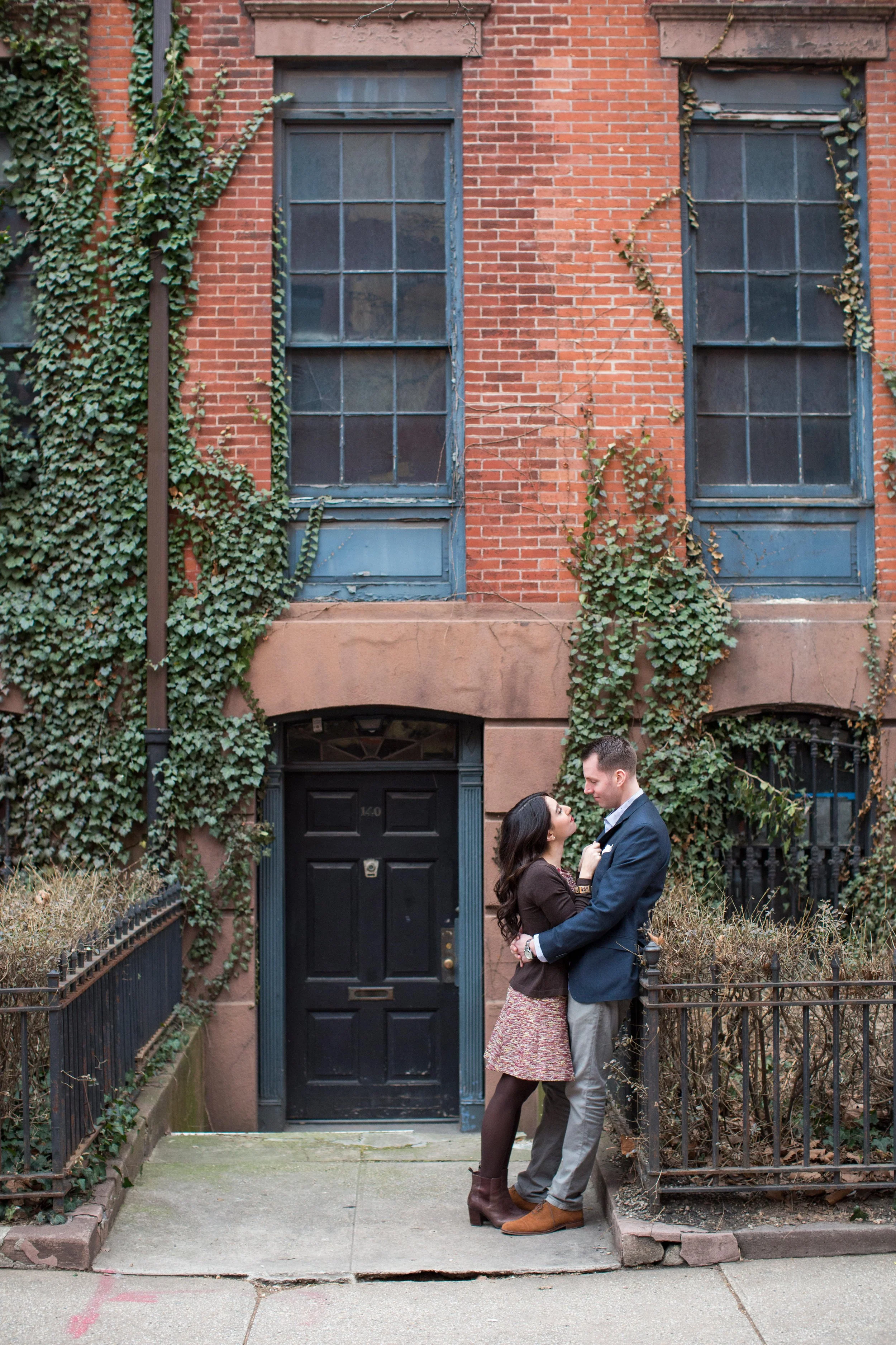 A couple stands close together on a city sidewalk in front of a brick building with ivy-covered walls and large multi-pane windows, sharing an intimate moment.