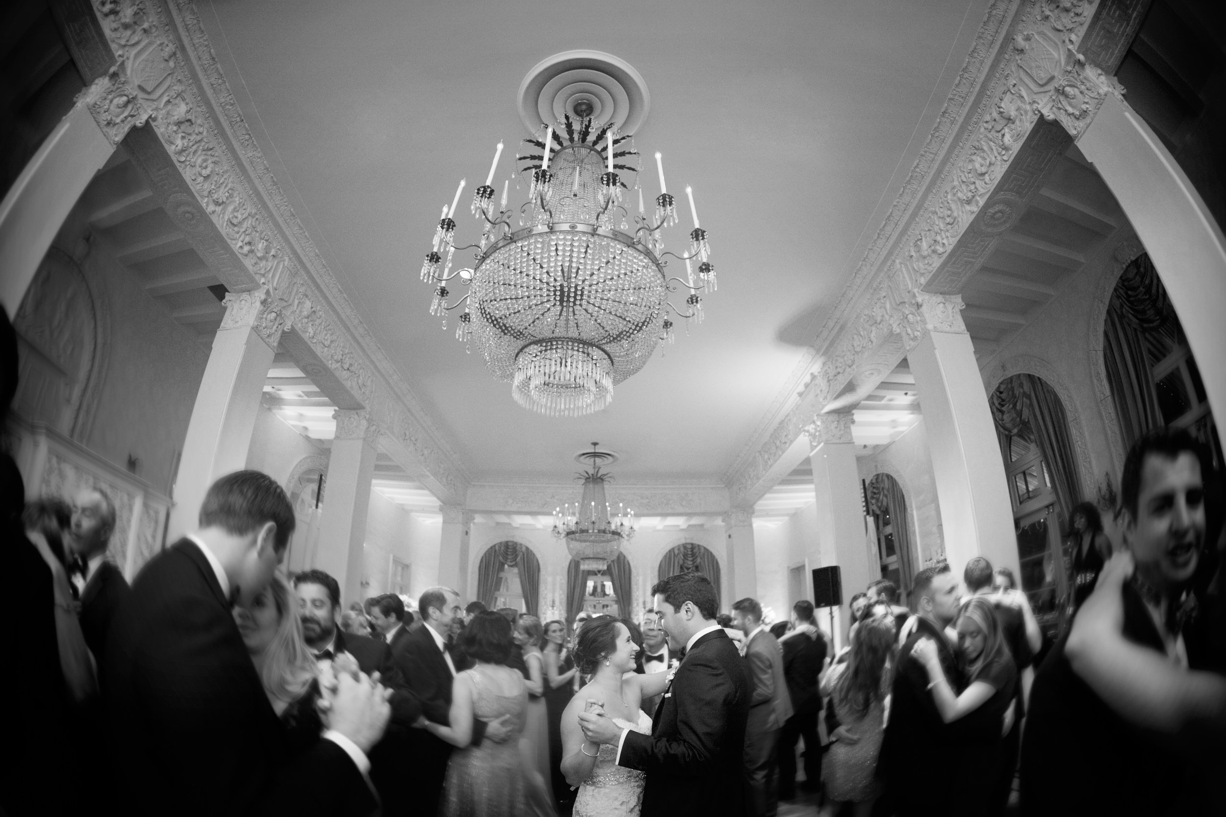 Black and white photo of a wedding reception with couples dancing in an elegant ballroom with ornate ceiling moldings and chandeliers.