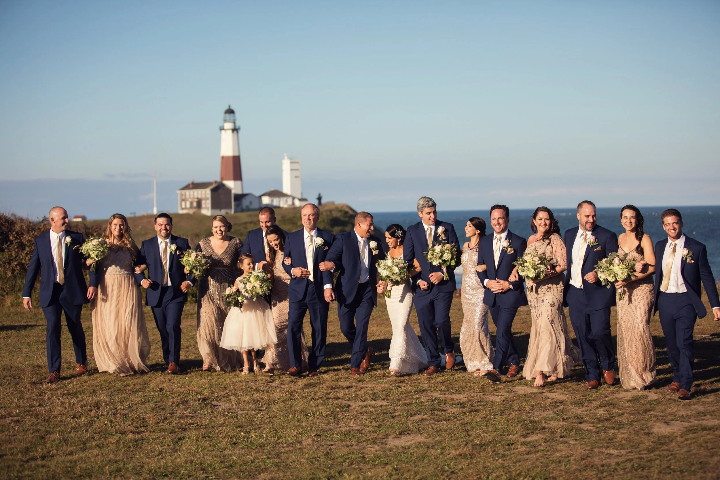 A group of wedding guests and the bride and groom walking arm in arm outdoors on a grassy area with a lighthouse in the background during daytime.