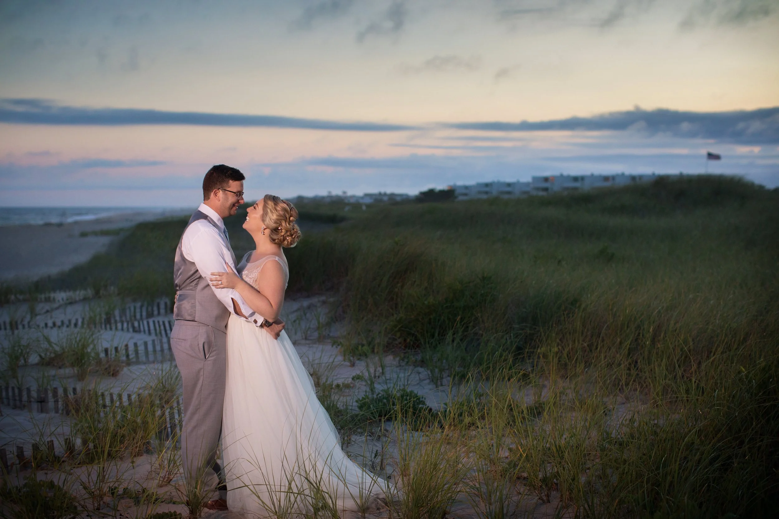 A couple in wedding attire happily embracing on a beach at sunset, with grassy dunes and a partly cloudy sky in the background.
