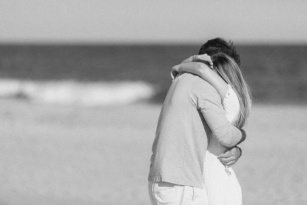 A black and white photo of a couple hugging on the beach, with the ocean in the background.