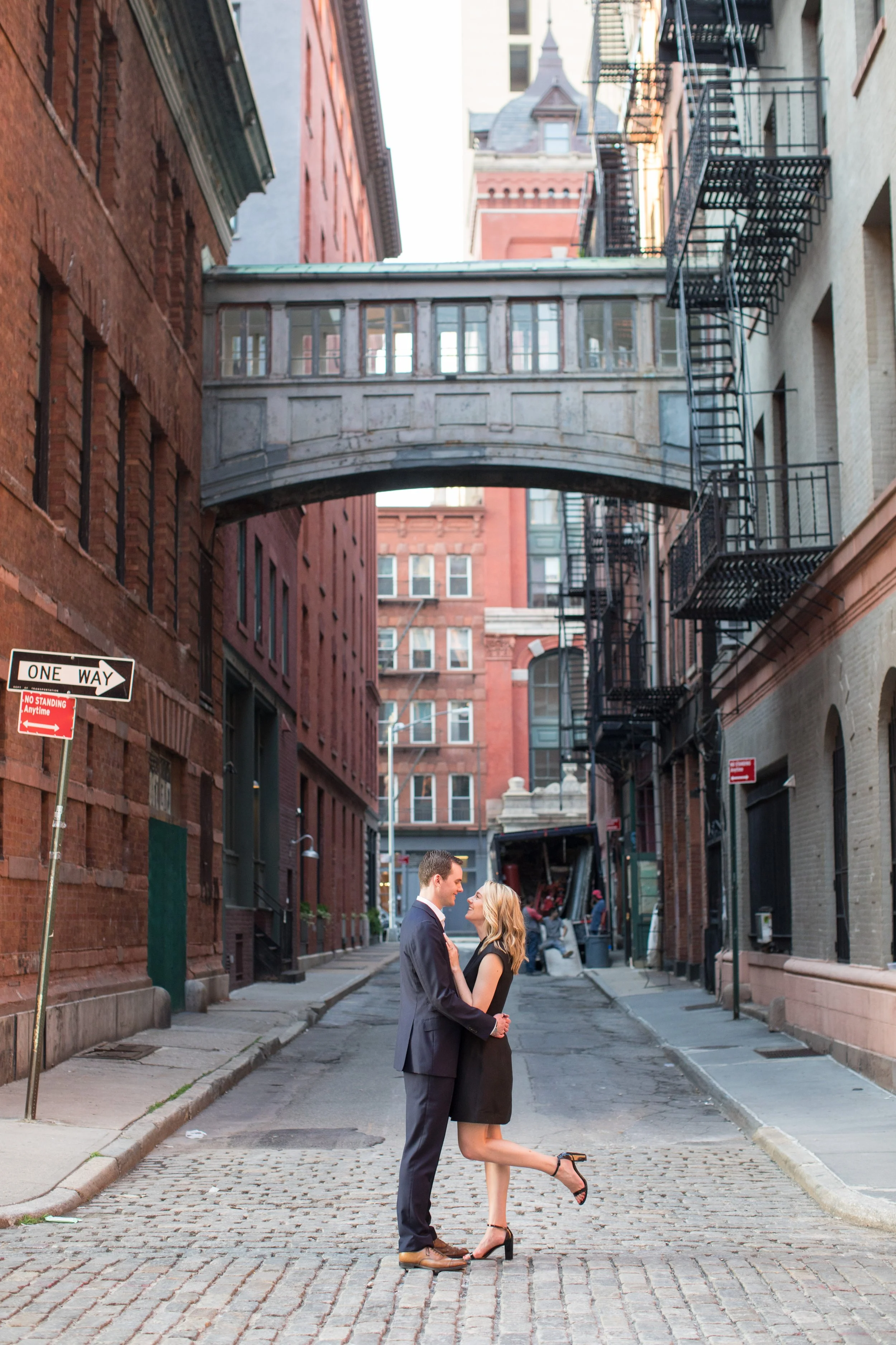 A couple in formal attire stands in a city alley beneath a bridge, sharing a romantic moment.