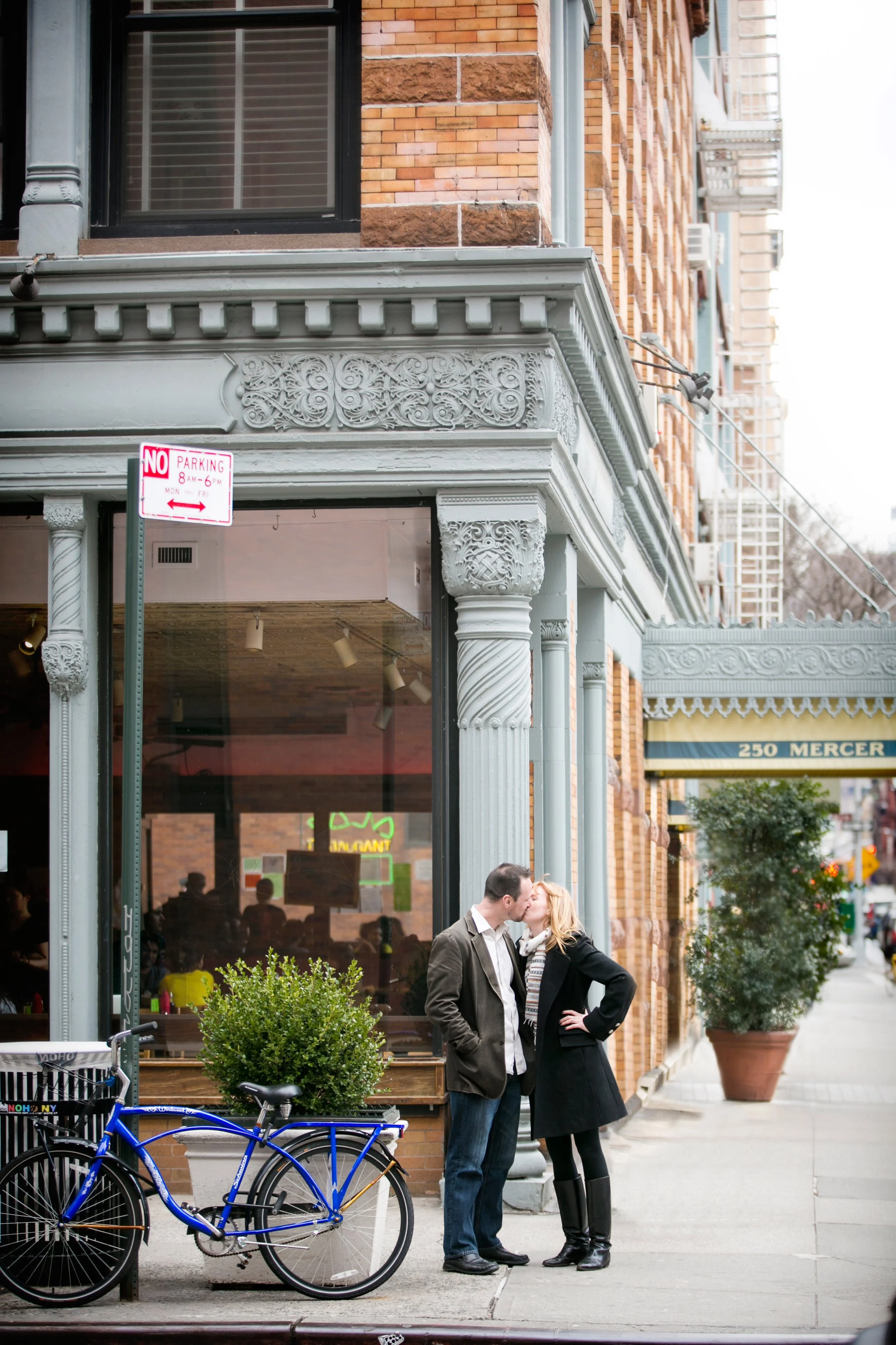 A couple kissing on a city sidewalk in front of a cafe with ornate architectural details, a large window, and a bicycle parked nearby.