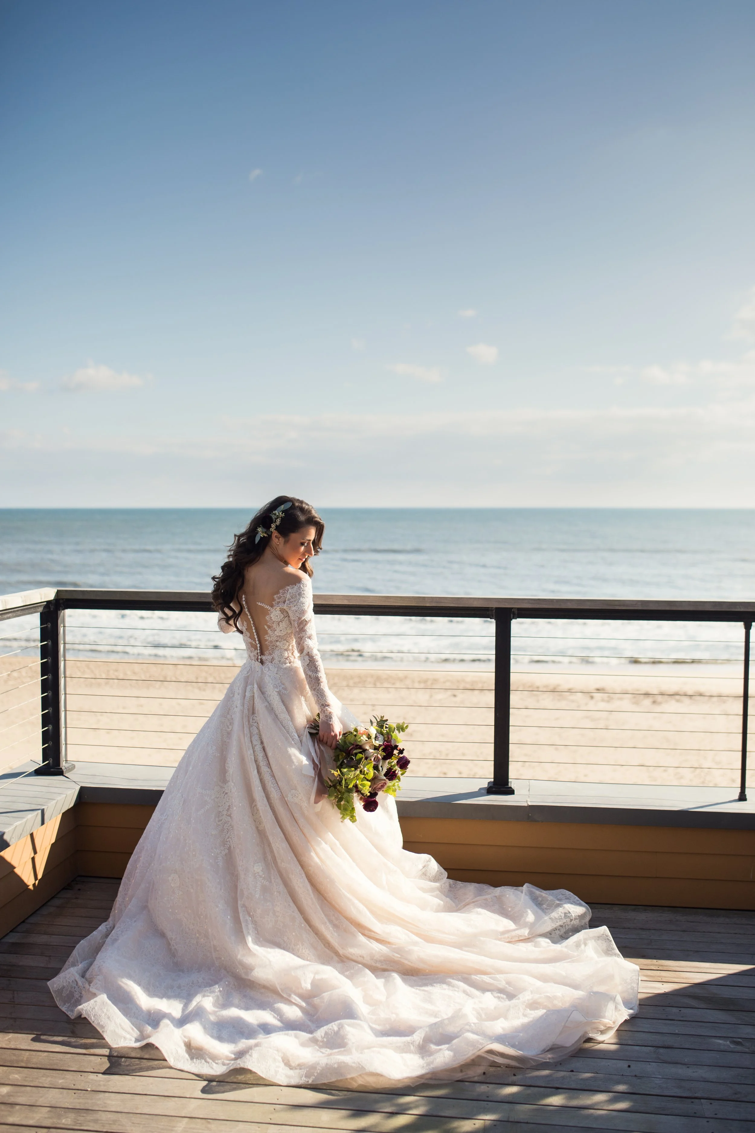 A bride in a white wedding dress holding a bouquet, standing on a wooden deck by the beach with the ocean and sky in the background.