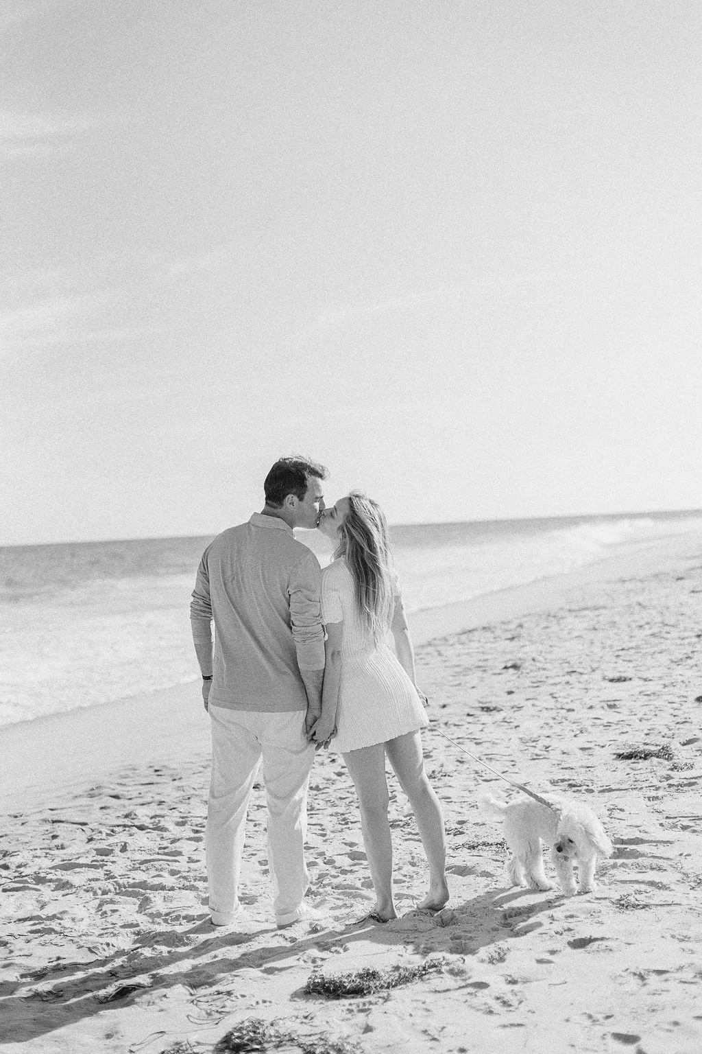 A couple kissing on the beach, holding hands, with a dog on a leash. The scene is in black and white.