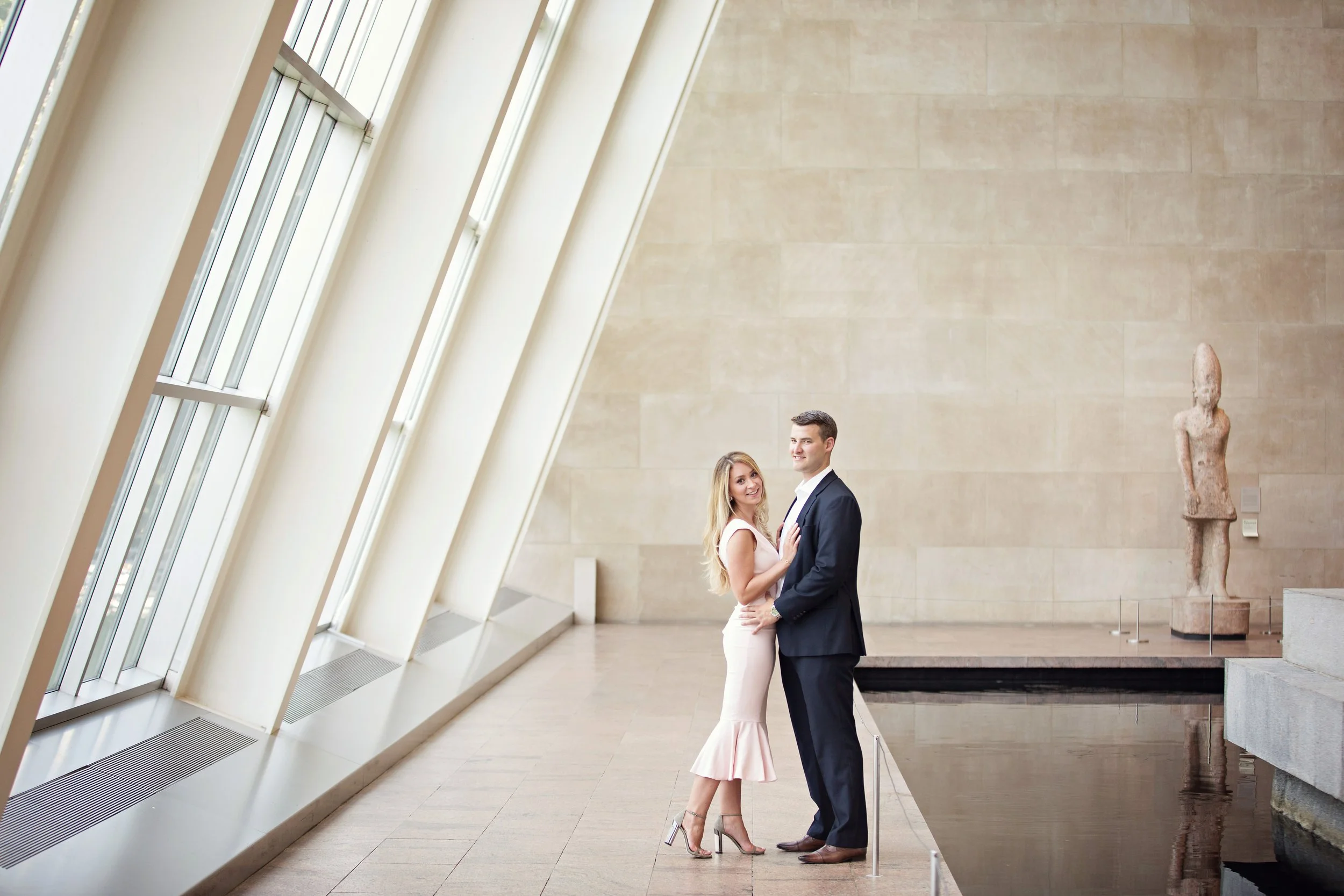 A smiling woman in a white dress and high heels and a man in a dark suit stand close together in an art gallery, with beige walls, large windows, and a sculpture in the background.