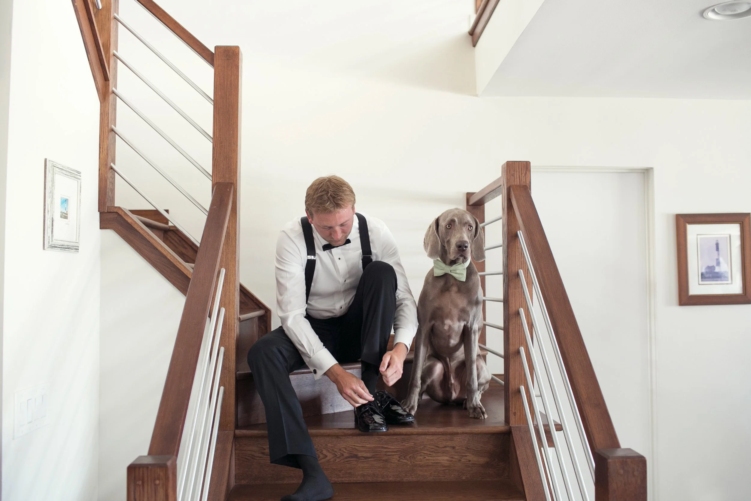 A man in formal attire with a bow tie is sitting on a staircase, tying his shoelaces. Next to him, a dog with a light-colored bow tie sits on the stairs, looking forward.