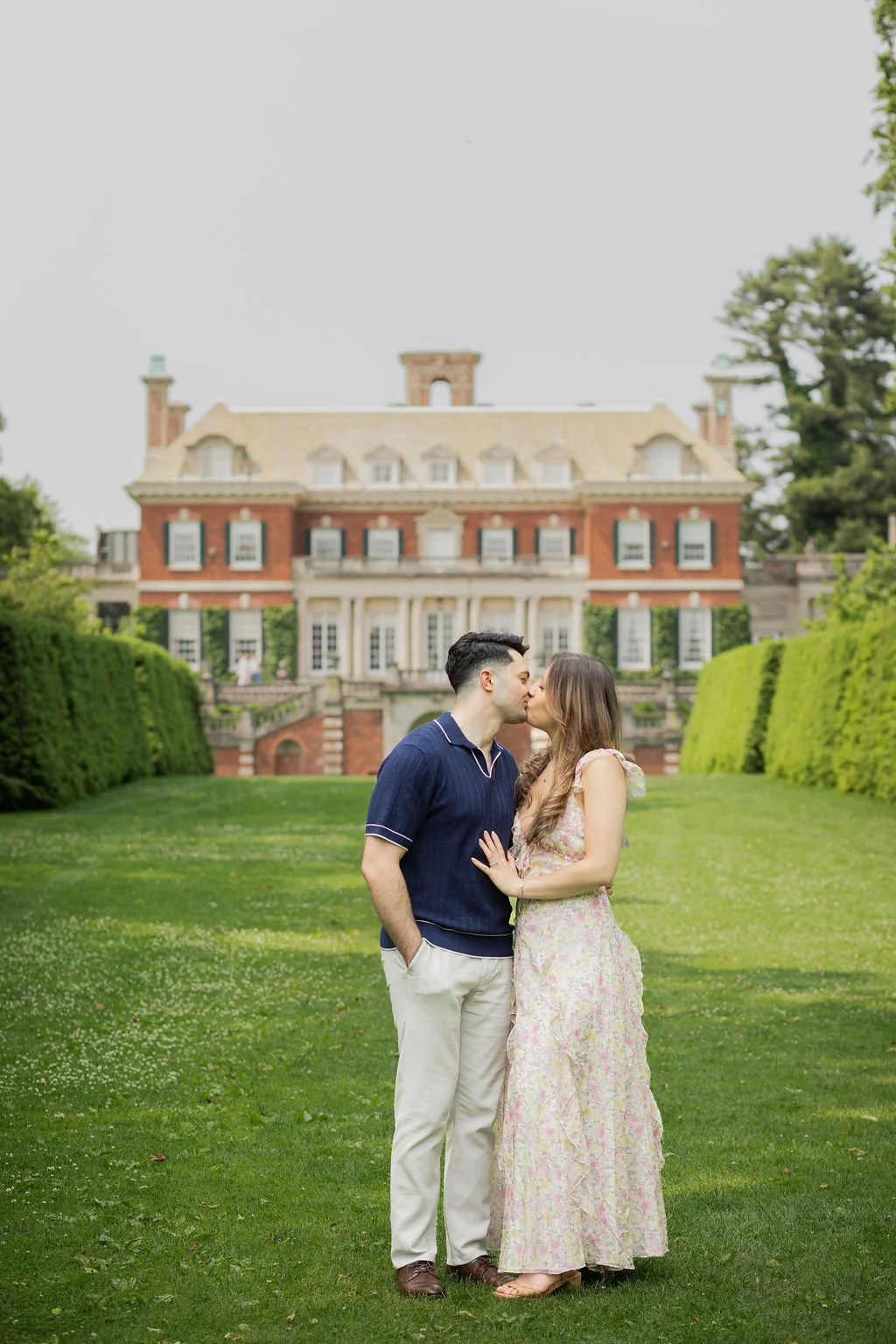 A couple kissing on a lush green lawn in front of a large historic mansion.