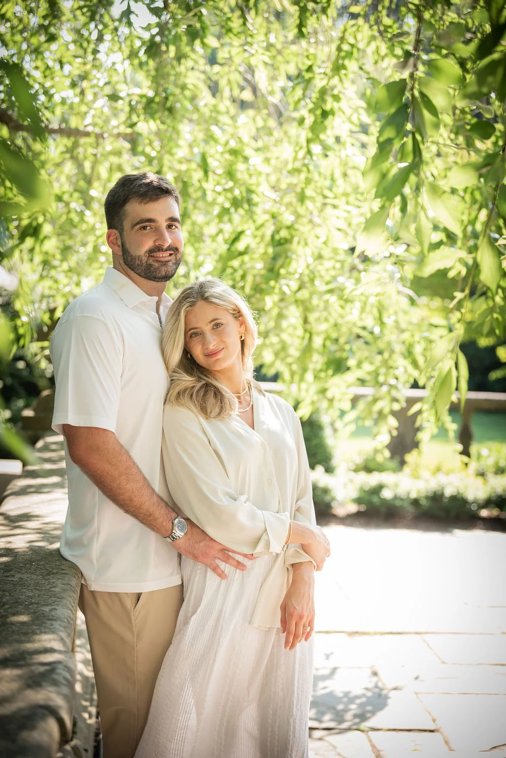A man and woman standing together outdoors amid green trees, with the man behind the woman, both smiling softly at the camera.