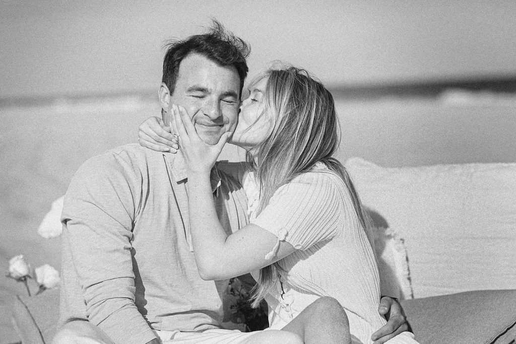 A woman kissing a man on the cheek at the beach, both smiling with eyes closed.