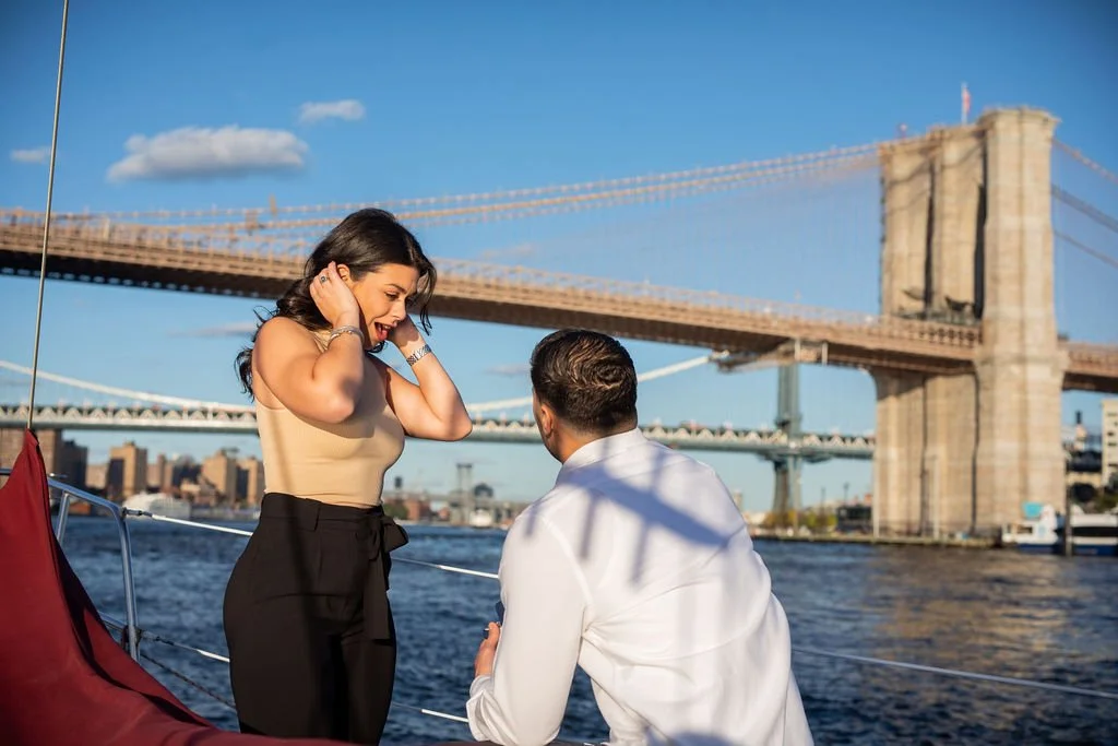 A woman standing on a boat talking to a man who is kneeling, with the Brooklyn Bridge and city skyline in the background.
