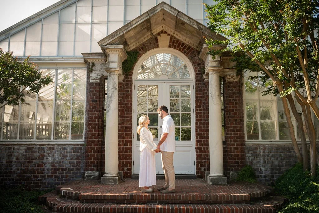 A couple holding hands and gazing at each other in front of a vintage brick greenhouse with white trim, surrounded by greenery.
