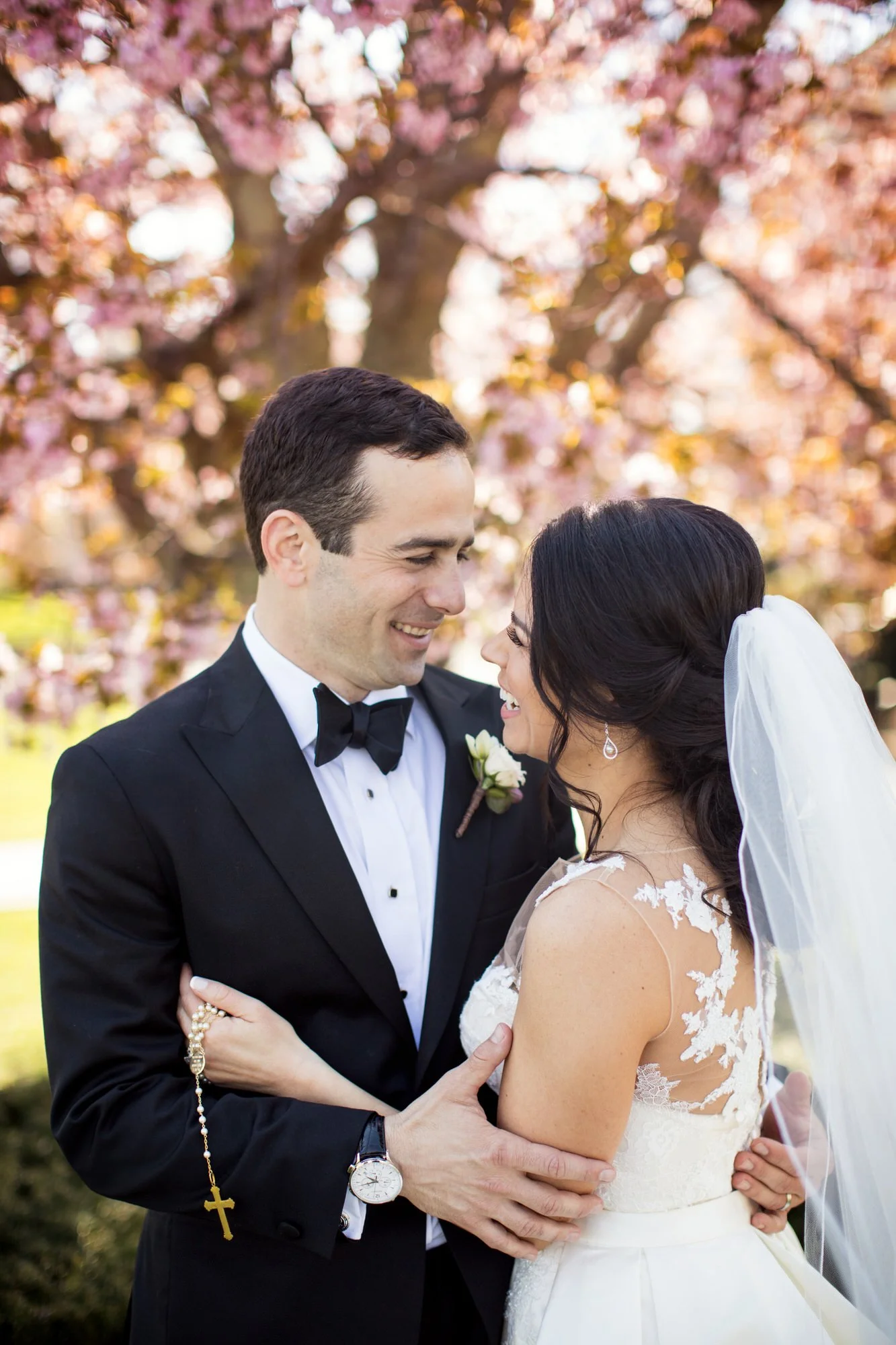 A groom and bride smiling at each other outdoors with pink blossom trees in the background.