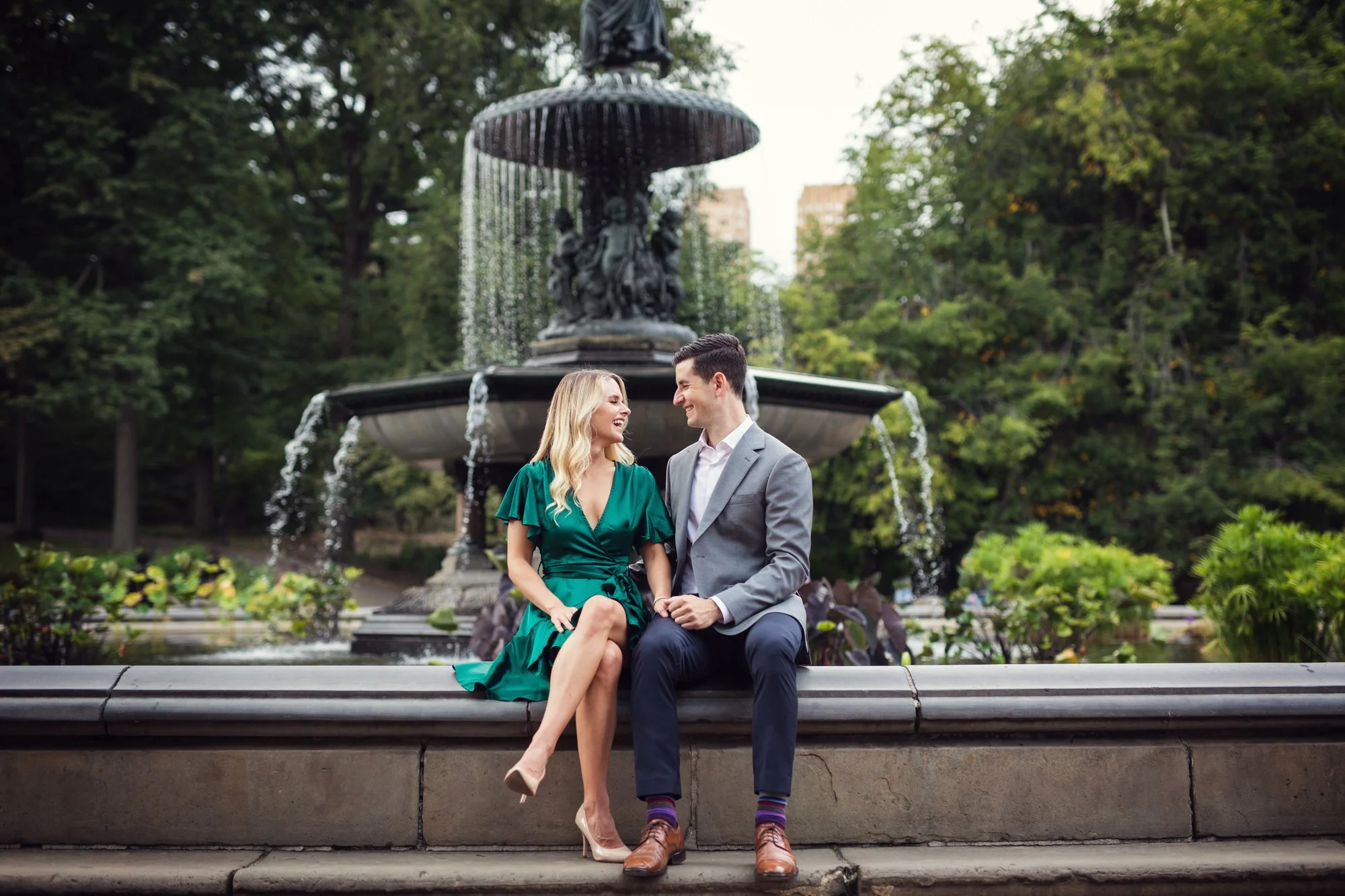A couple sitting on a park ledge in front of a large fountain with water cascading from it, surrounded by greenery.