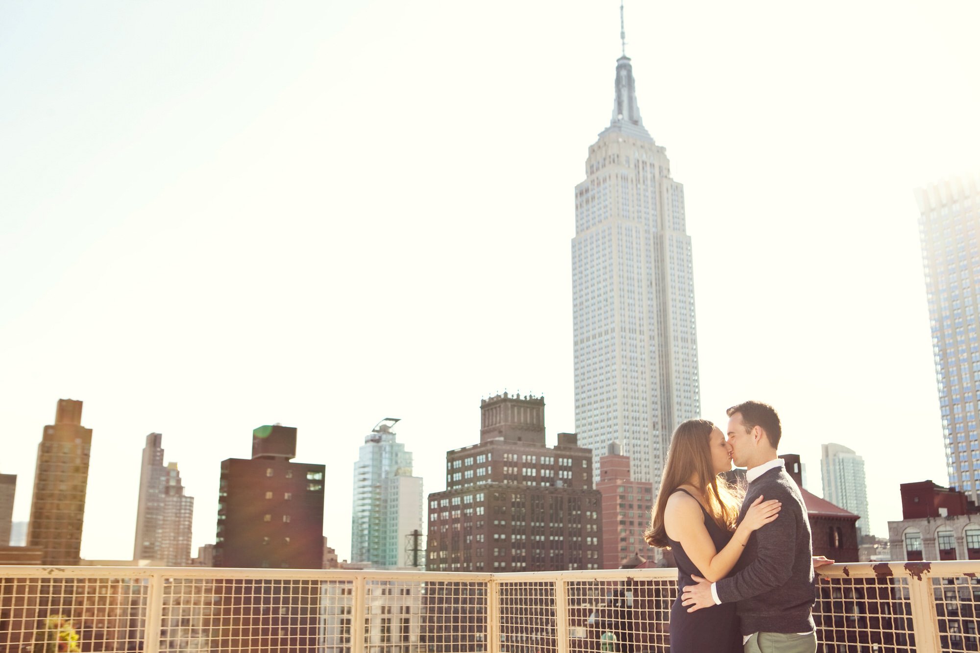 A couple kissing on a rooftop with the New York City skyline, including the Empire State Building, in the background.
