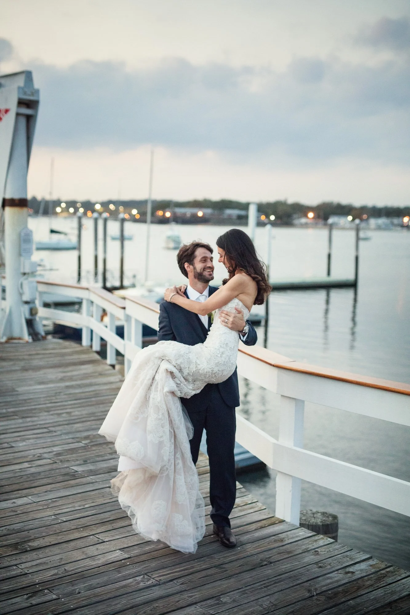 A groom lifting a bride on a dock by the water at sunset, with boats and sailboats in the background.