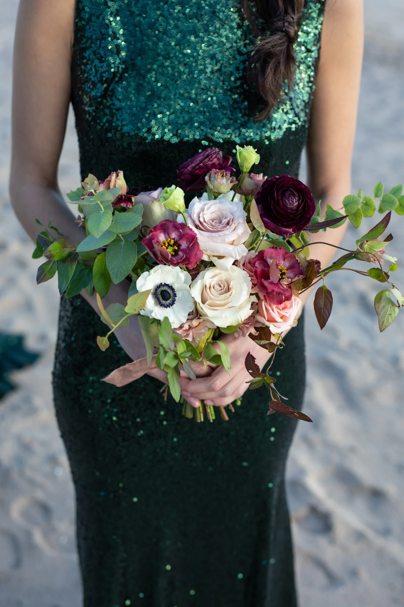 A woman in a dark green, sequined dress holding a colorful bouquet of flowers in both hands, standing outdoors on sandy ground.