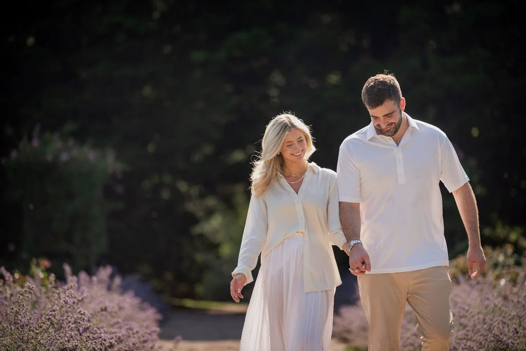A smiling couple walking hand in hand through a garden or park, dressed in light-colored clothing.