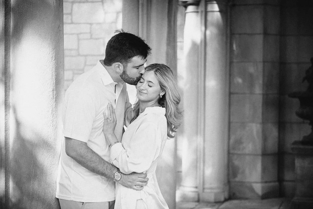 A couple embraces and shares an intimate moment in black and white, standing against a wall with architectural columns in the background.
