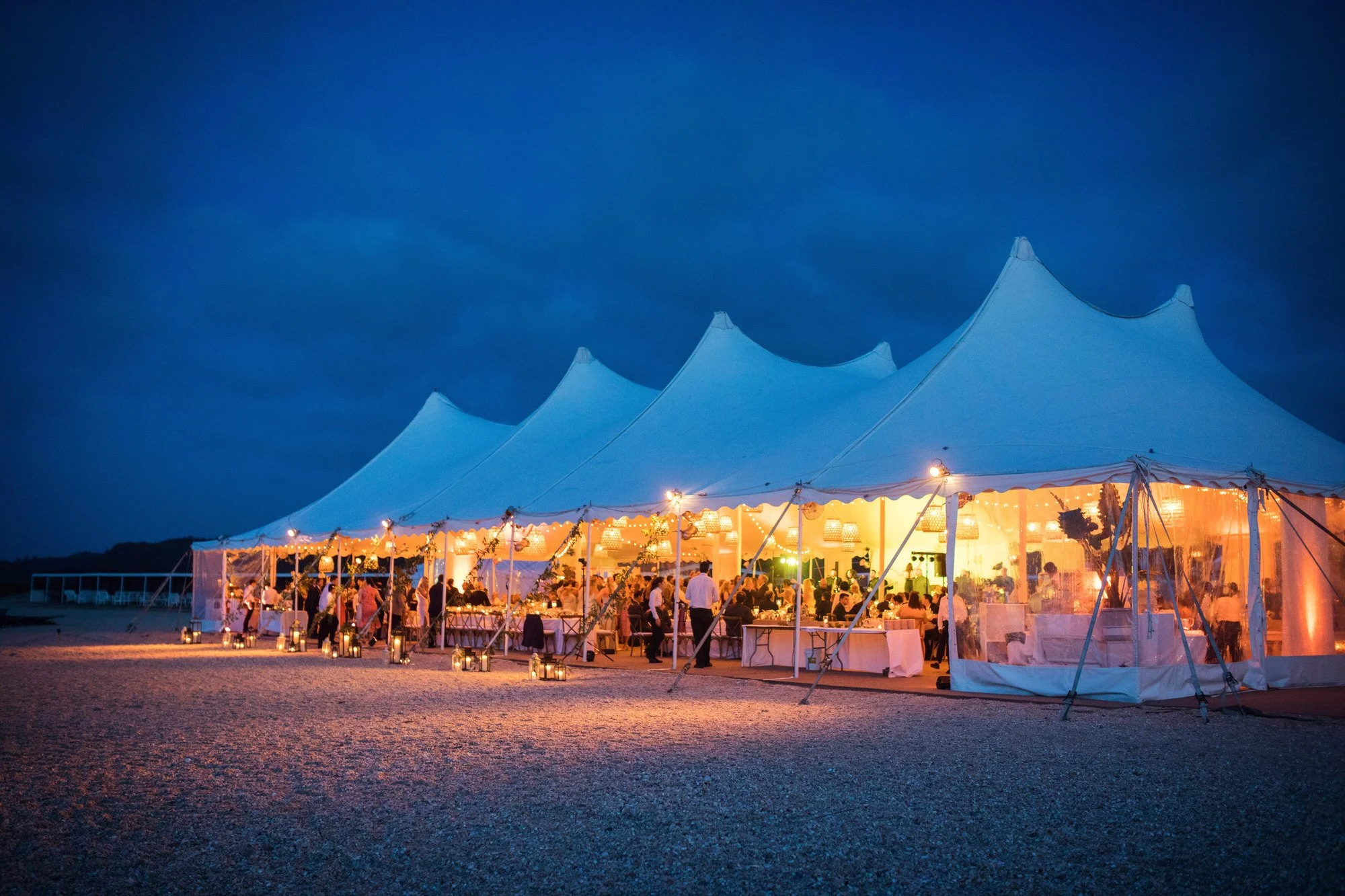 A large white event tent illuminated with warm string lights at night, set up on a gravel surface with tables and guests inside, against a dark blue sky.