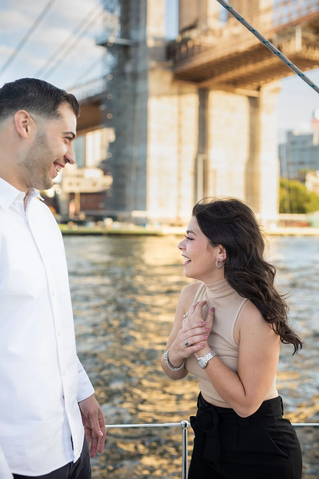 A man and woman smiling and laughing near a river with a bridge in the background.