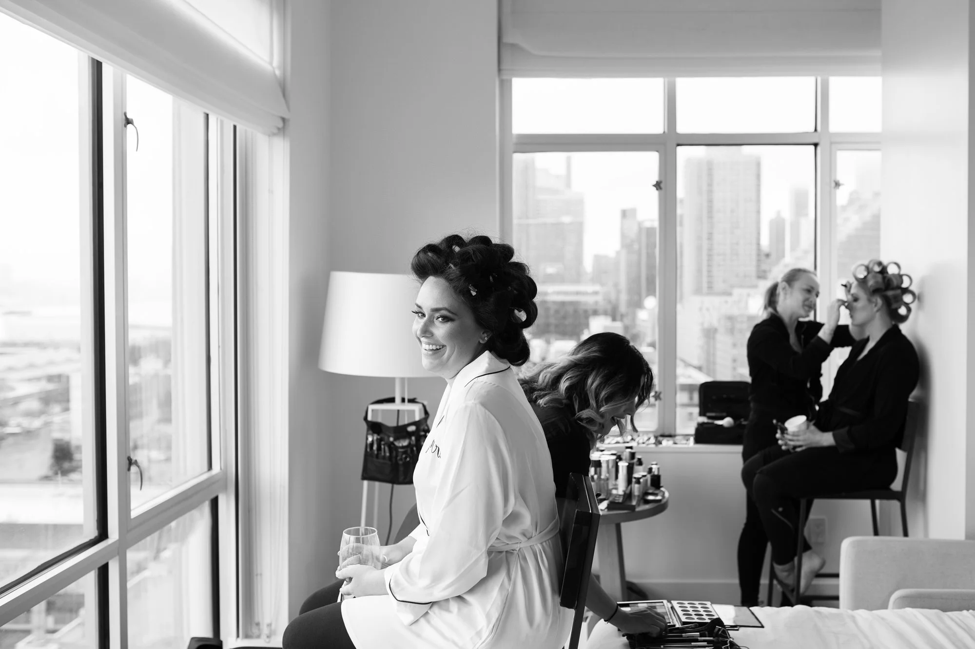 Women preparing for a wedding in a city apartment, with one woman getting her makeup done in the background, all women with hair rollers, a smiling bride holding a glass while sitting near a large window.