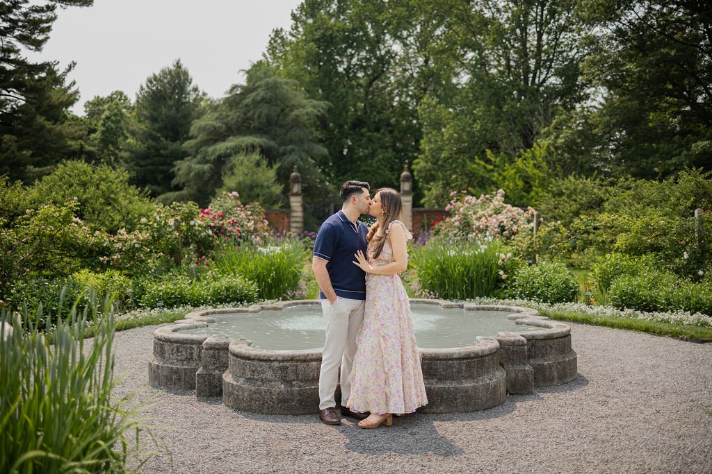 A couple stands close, about to kiss, in front of a stone fountain in a lush, flowering garden.