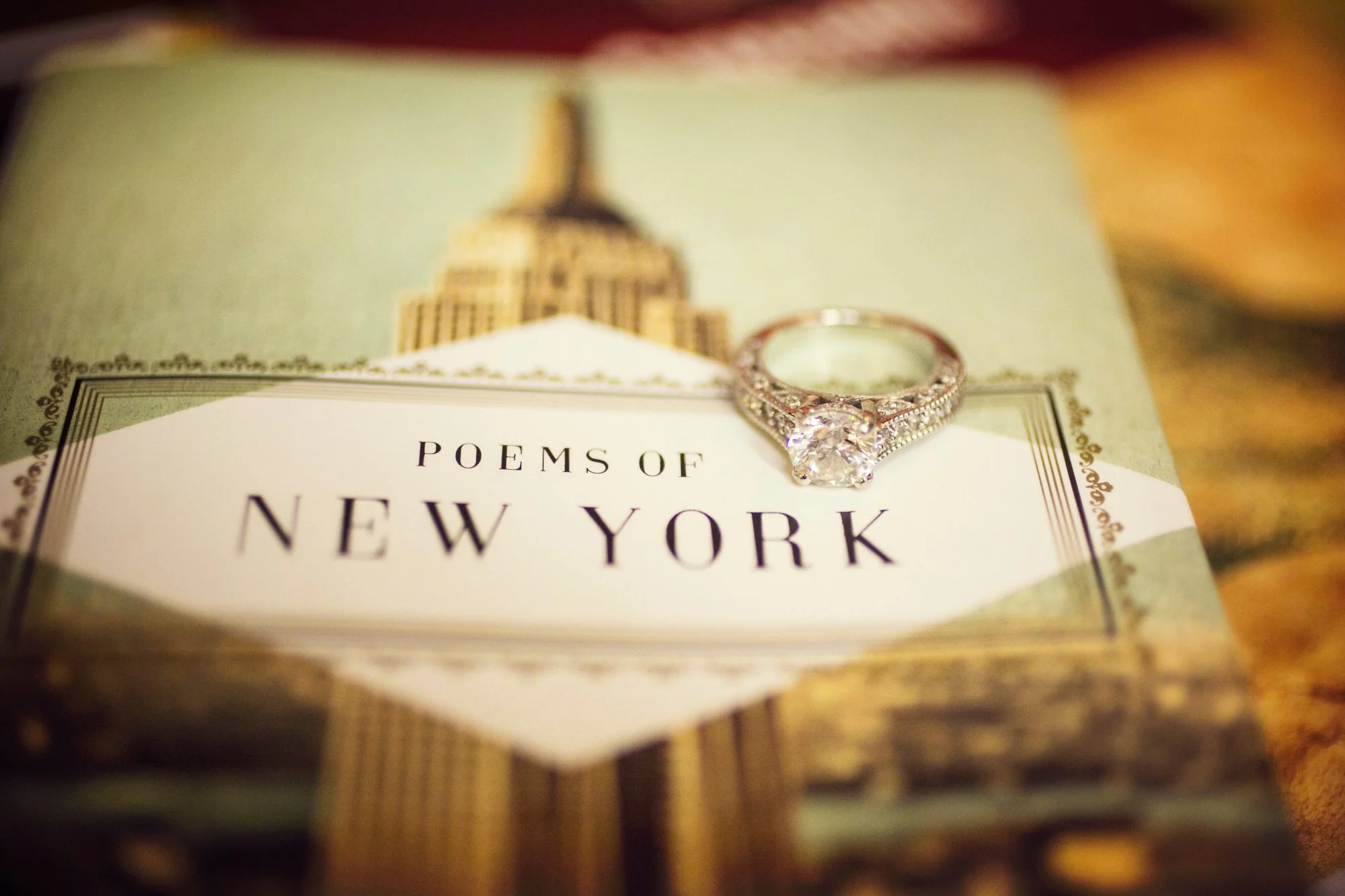 Close-up of a book titled 'Poems of New York' with a diamond engagement ring resting on it, and an illustration of a New York City building in the background.