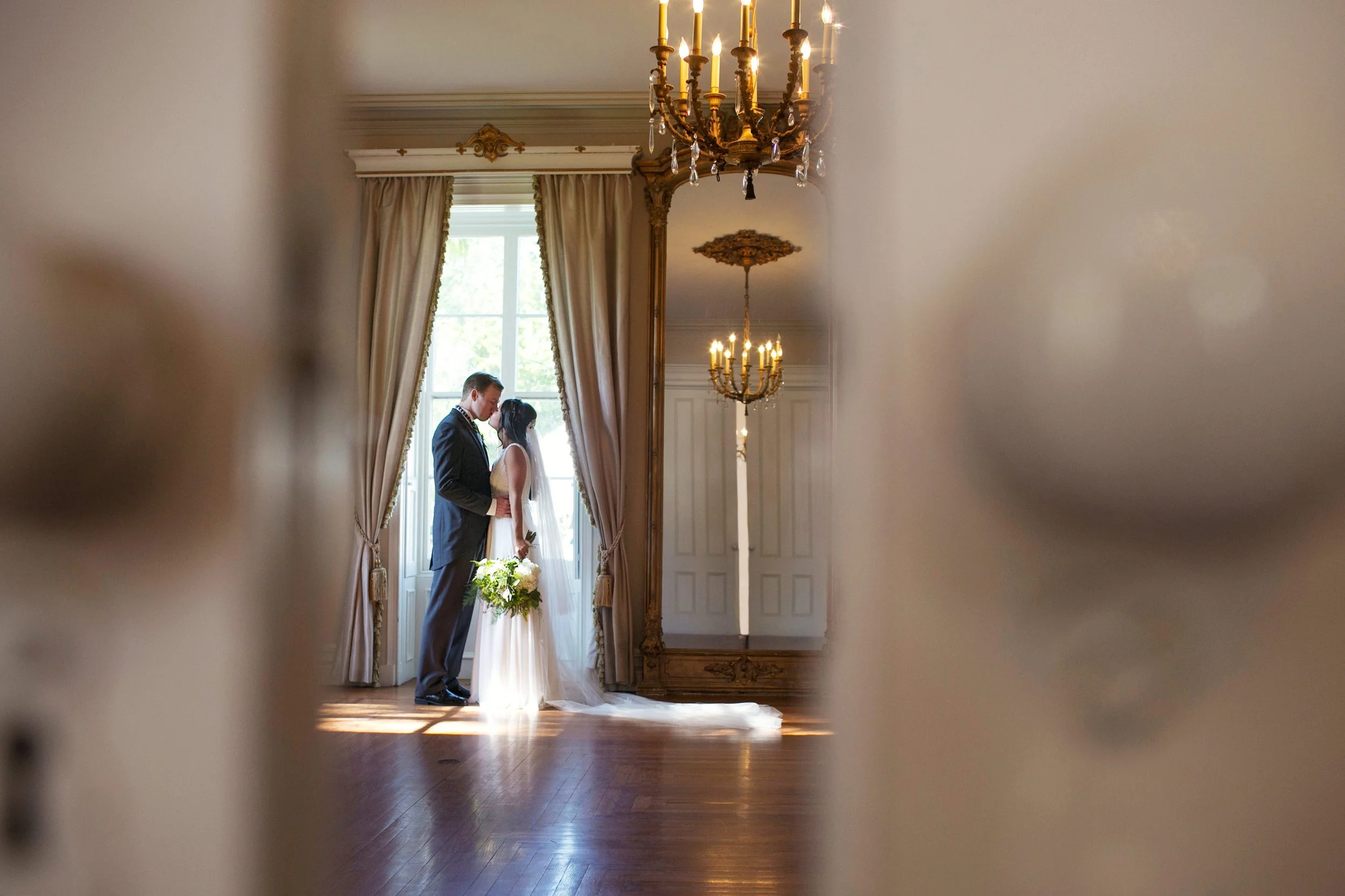 A bride and groom standing close together, holding hands, with a large window and curtains behind them, inside an elegant room with chandeliers and wooden floors, seen through an opening.