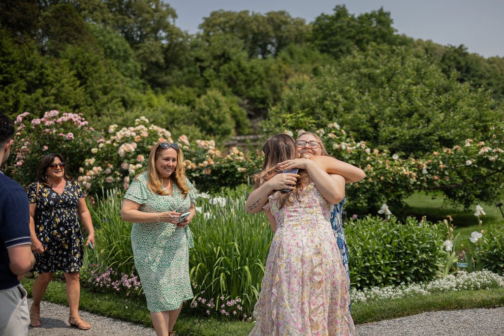 Two women hugging and smiling in a garden surrounded by flowers, with three other women nearby and lush green trees in the background.