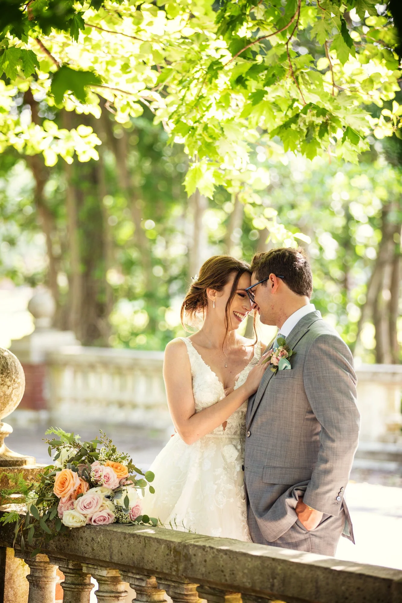 A bride and groom sharing a romantic moment outdoors in a garden with green trees overhead, dressed in wedding attire, with a bouquet of flowers on the stone railing nearby.