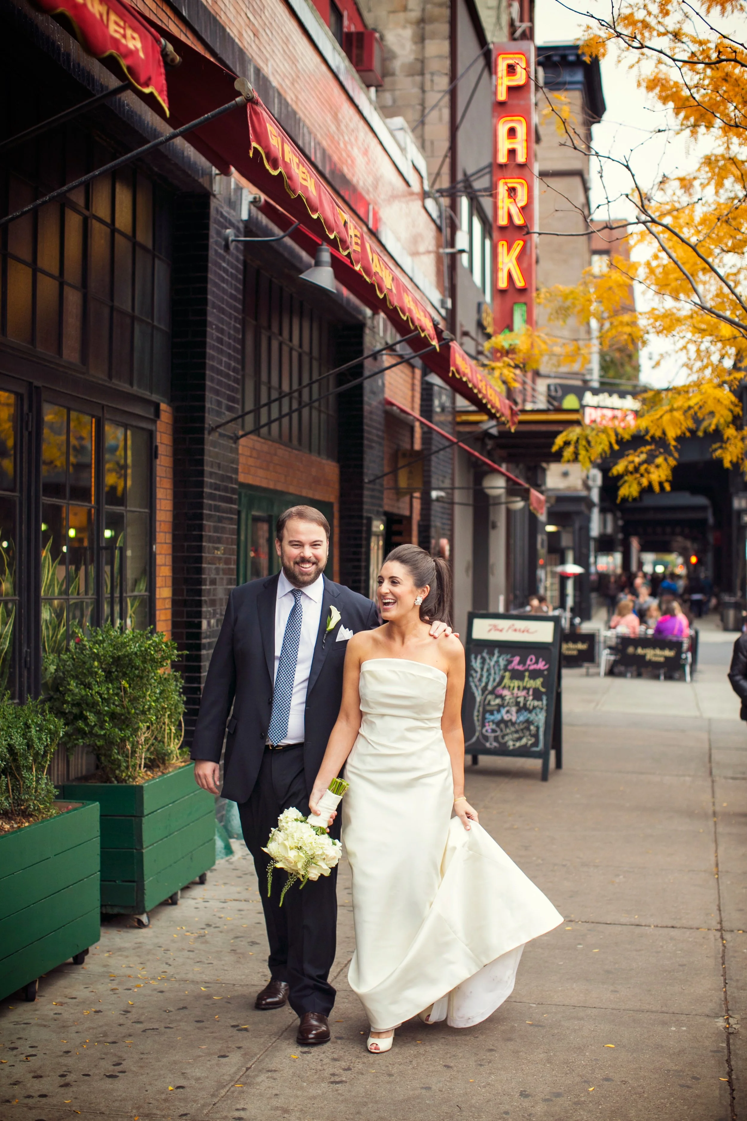 A newlywed couple walking on a city sidewalk, smiling and holding a bouquet of white flowers, with a brick building and neon park sign in the background.