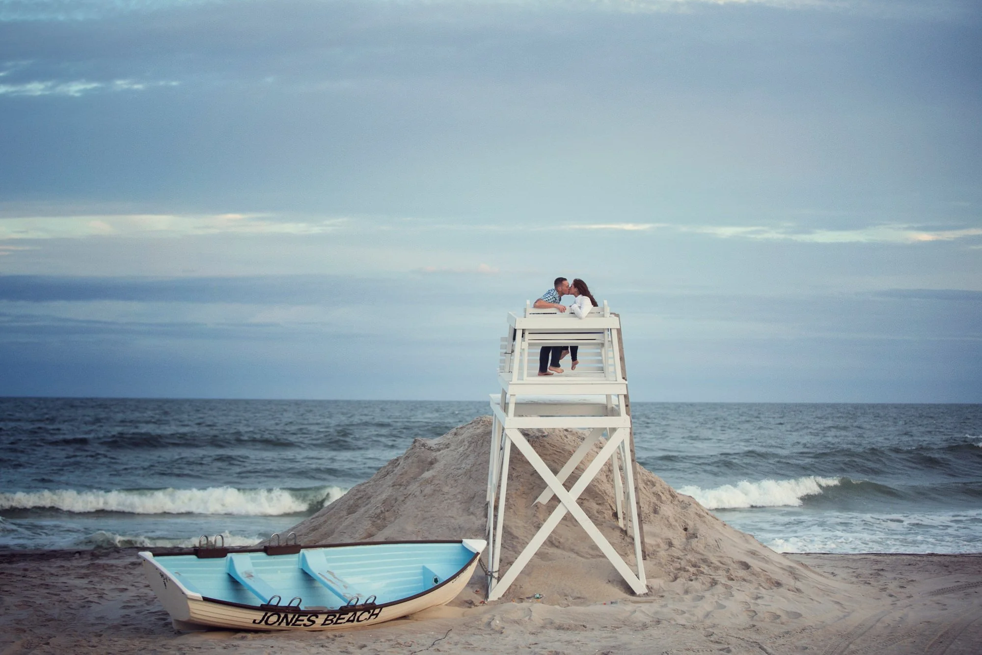 A couple kisses on a white lifeguard tower on the beach with a blue boat labeled 'Jones Beach' on sandy shore and ocean waves in the background.