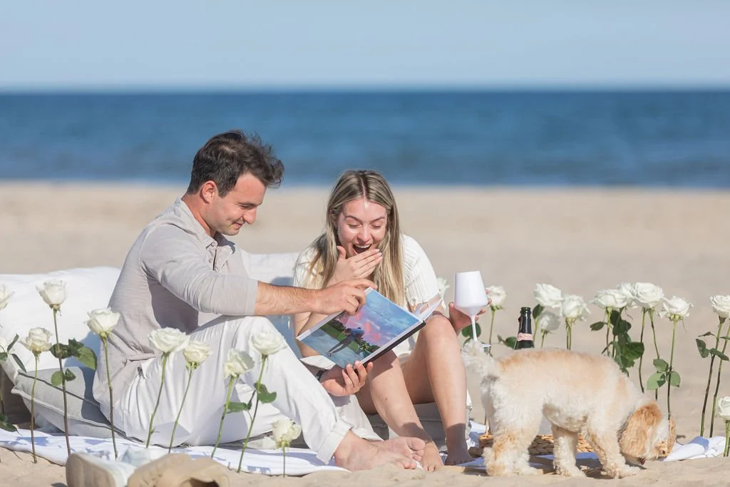 A couple sitting on a white blanket at the beach, looking at a photo album and laughing, with white roses surrounding them, a dog sniffing the sand, and the ocean in the background.