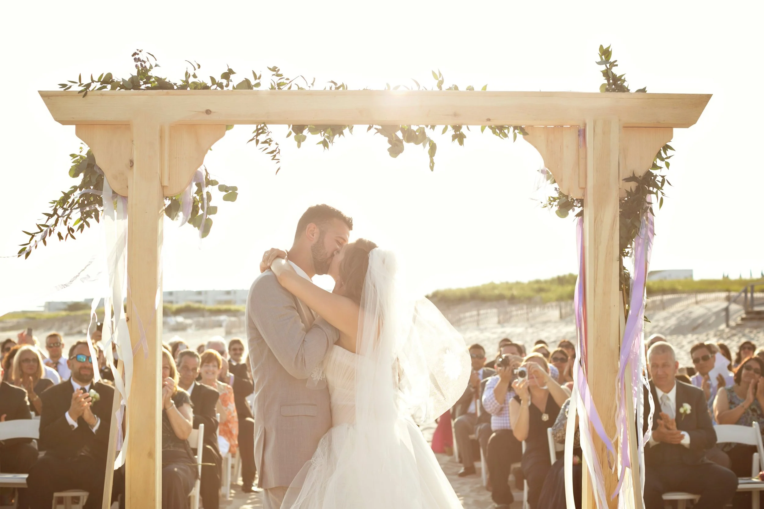 A couple sharing a kiss during their wedding ceremony on the beach, surrounded by seated guests with some taking photos, under a wooden arch decorated with greenery and purple ribbons at sunset.