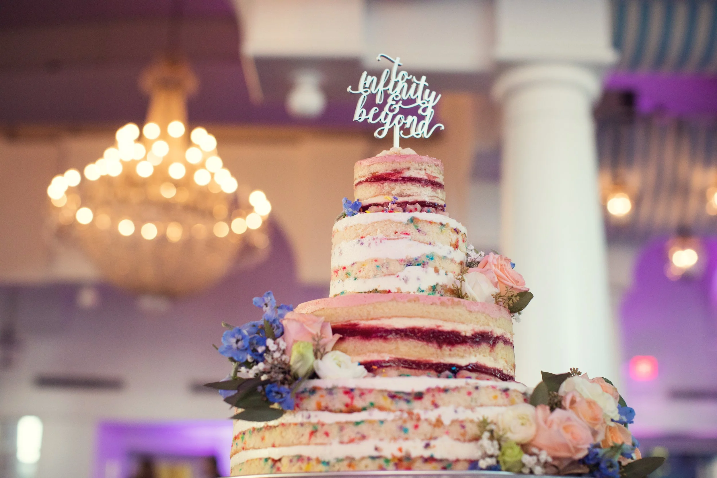 A tiered cake with semi-naked frosting and colorful sprinkles, decorated with pink, peach, and white roses along with blue flowers, topped with a cake topper that says 'to infinity & beyond,' in a decorated reception hall with a chandelier in the bac