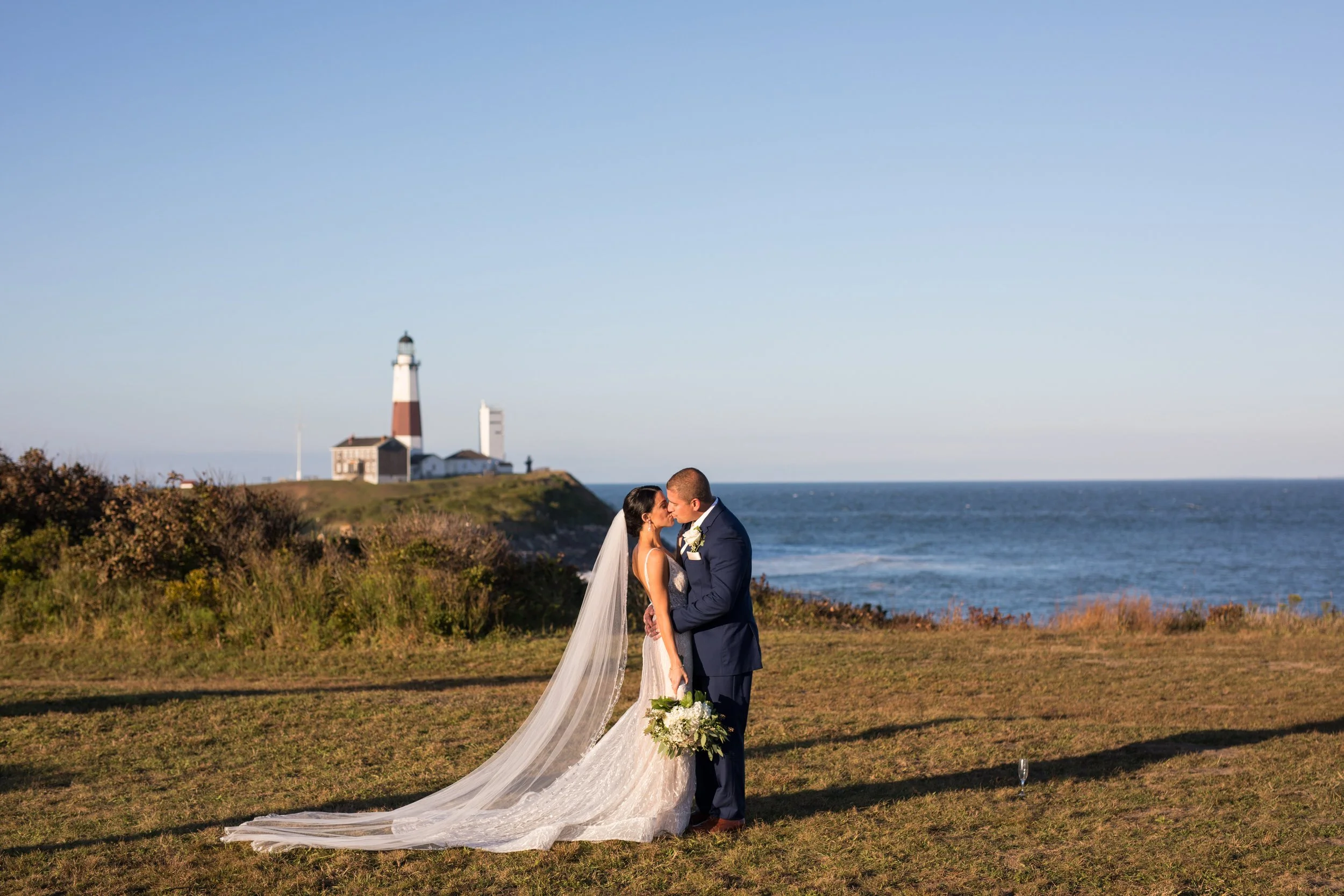 A bride and groom kissing outdoors near the ocean with a lighthouse in the background.