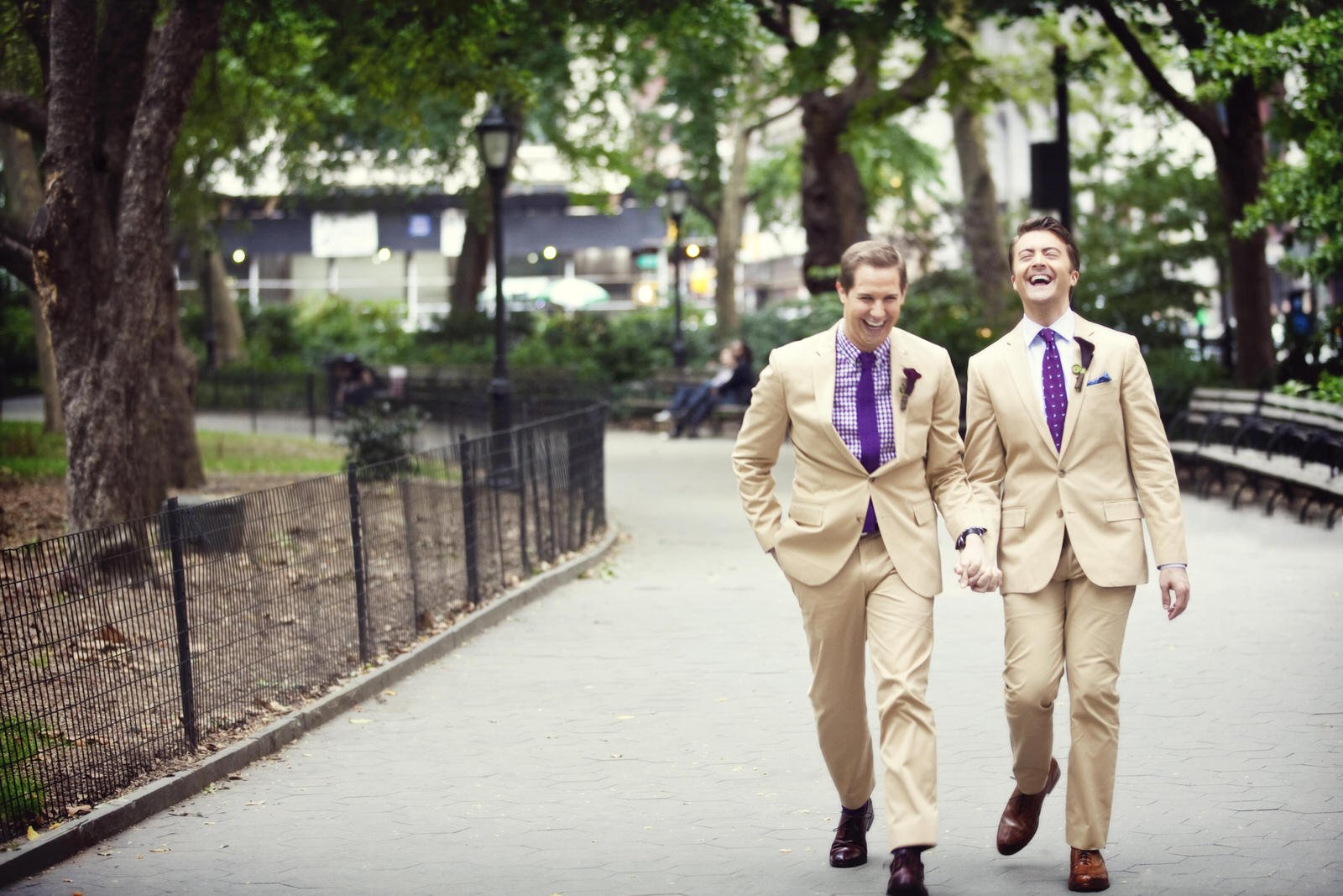 Two men in beige suits walking in a park, holding hands and laughing.