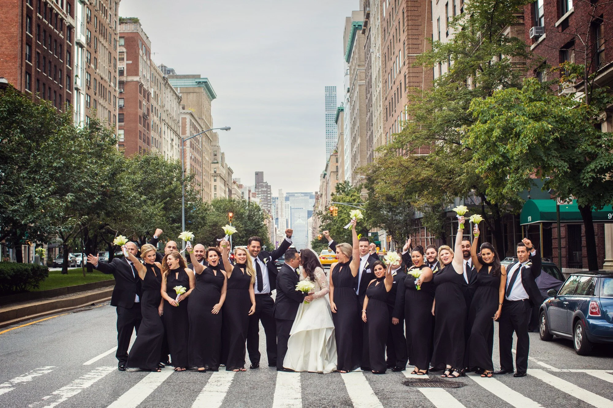 A wedding party standing in the middle of a city street crossing, celebrating and holding white flowers, with tall buildings and trees in the background.