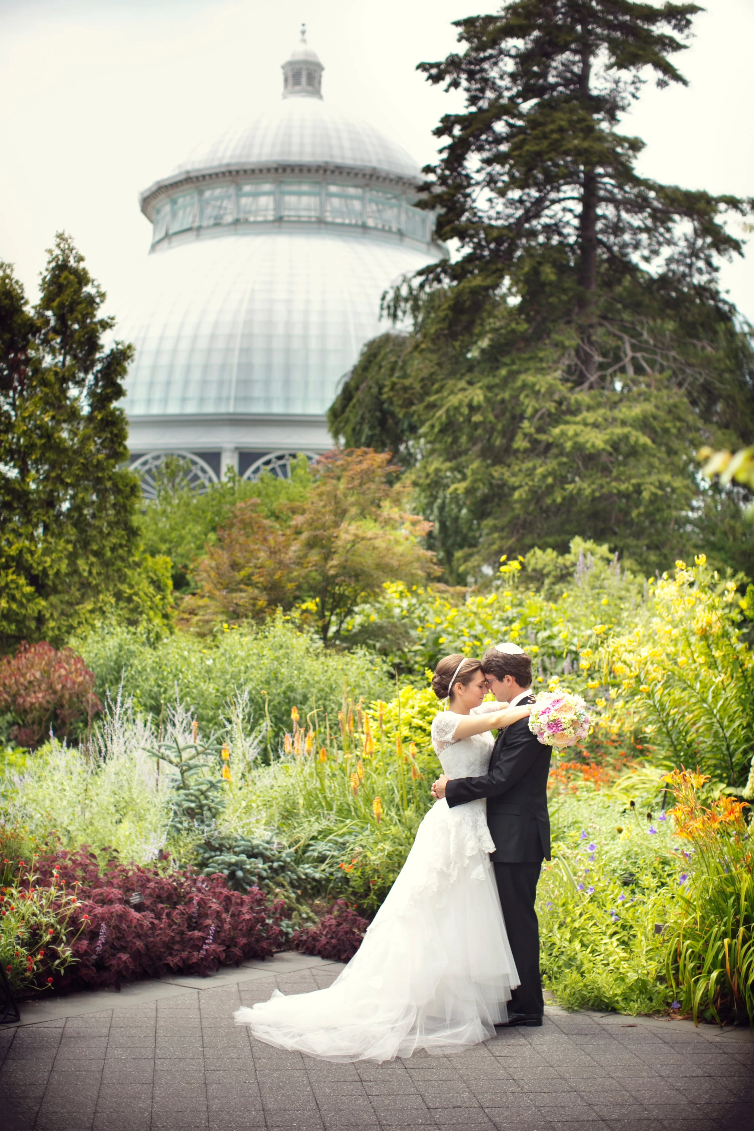 A bride and groom embrace in a garden with colorful flowers, a large domed building in the background, and tall trees around.