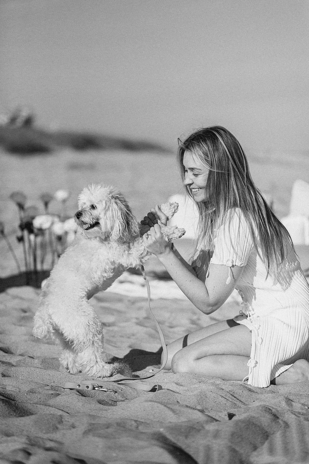 A woman and a dog playing together on the beach, with the woman kneeling on the sand and the dog standing on its hind legs, holding the woman's hands. The woman has long hair and is smiling, while the dog looks happy. The scene is set on a sandy beac