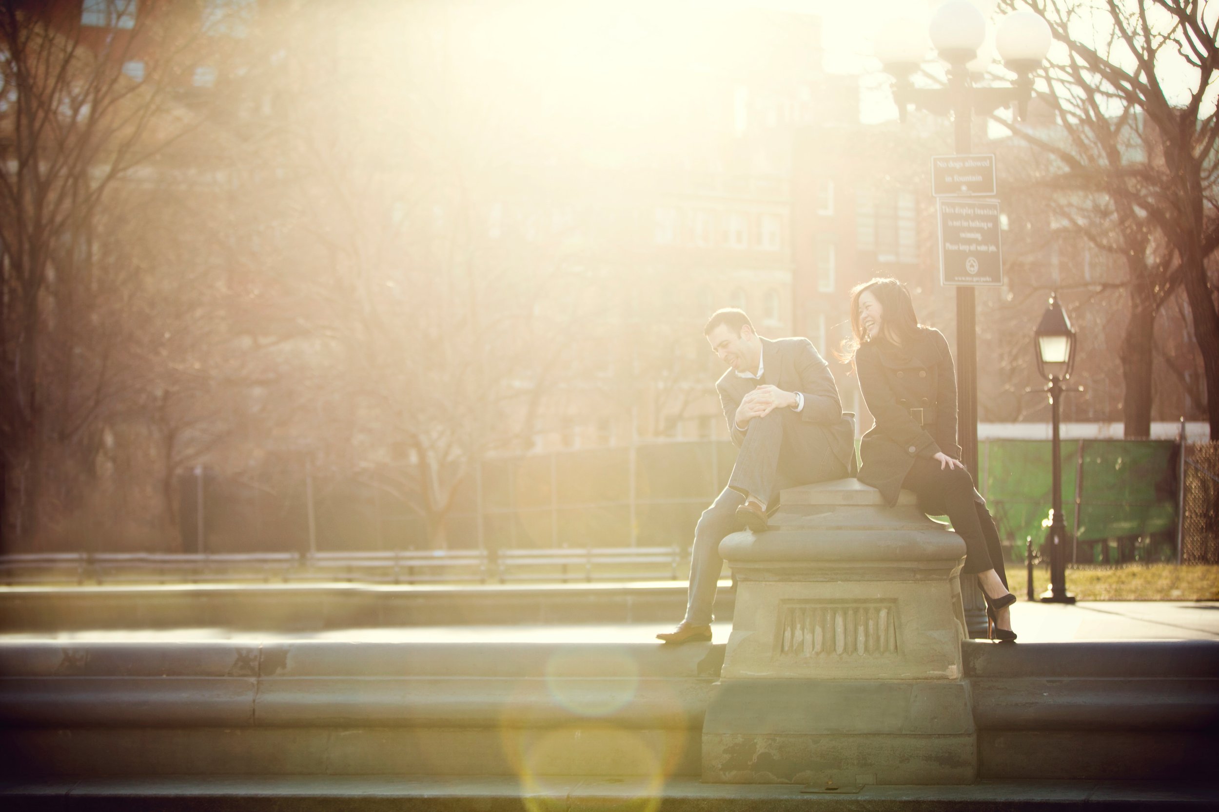 A man and woman sitting on a park bench, smiling and laughing, with sunlight shining brightly behind them.