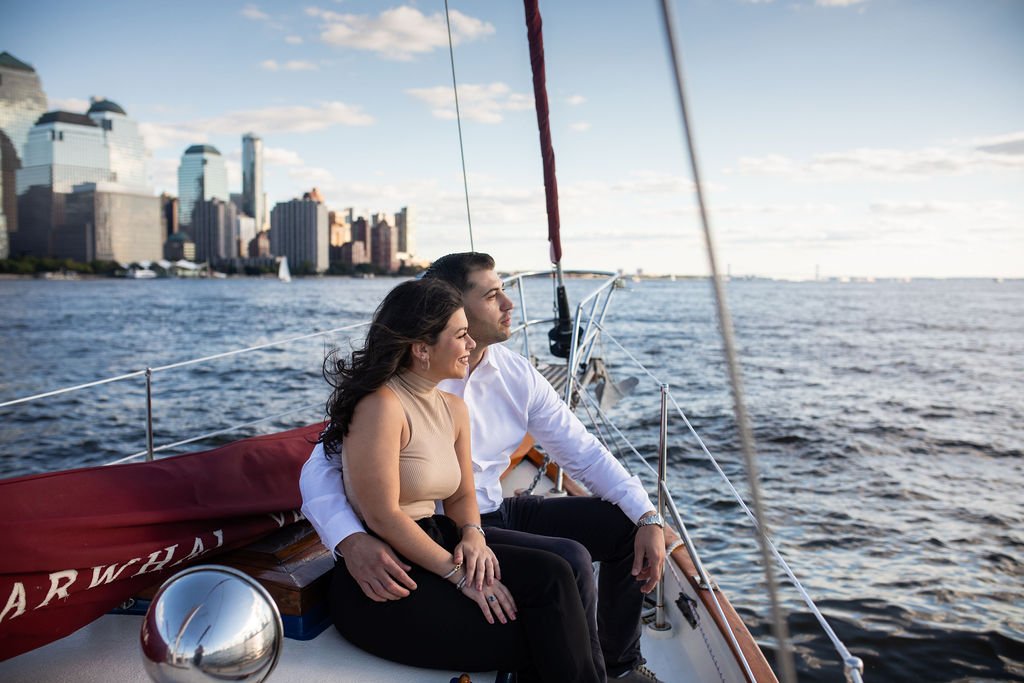 A young couple sitting on a sailboat with a city skyline in the background, enjoying a moment on the water.