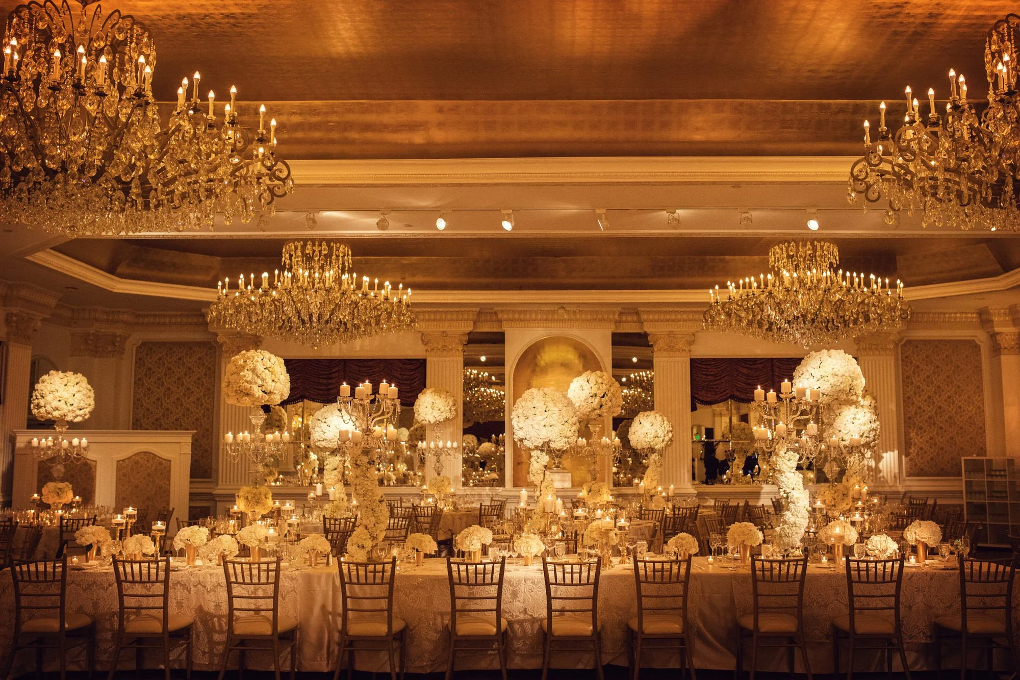 Elegant banquet hall decorated with numerous white floral centerpieces, candelabras, and luxurious chandeliers, set for a formal event.