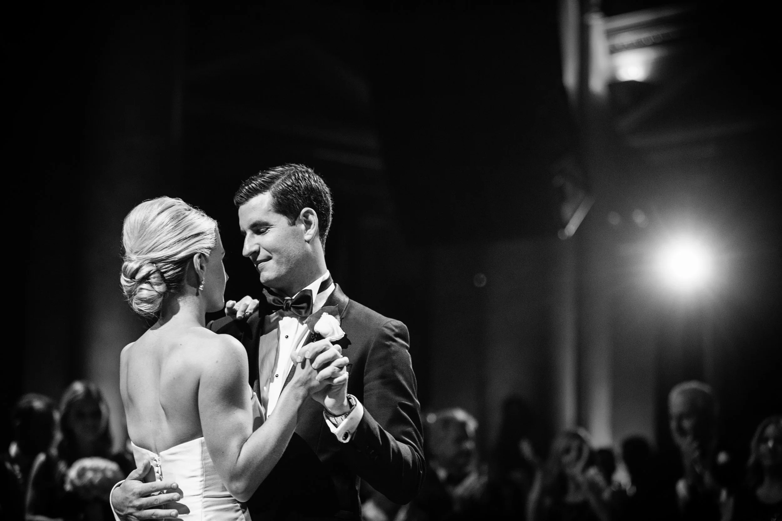 A black and white photograph of a bride and groom dancing, with the bride in a strapless white dress and the groom in a tuxedo, at their wedding reception.