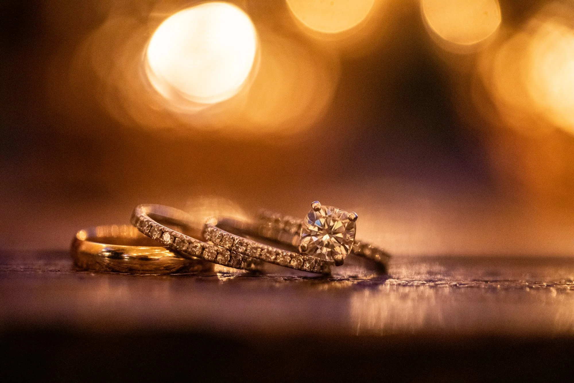 Close-up of three wedding rings, one with a large diamond, set on a reflective surface with blurred golden lights in the background.
