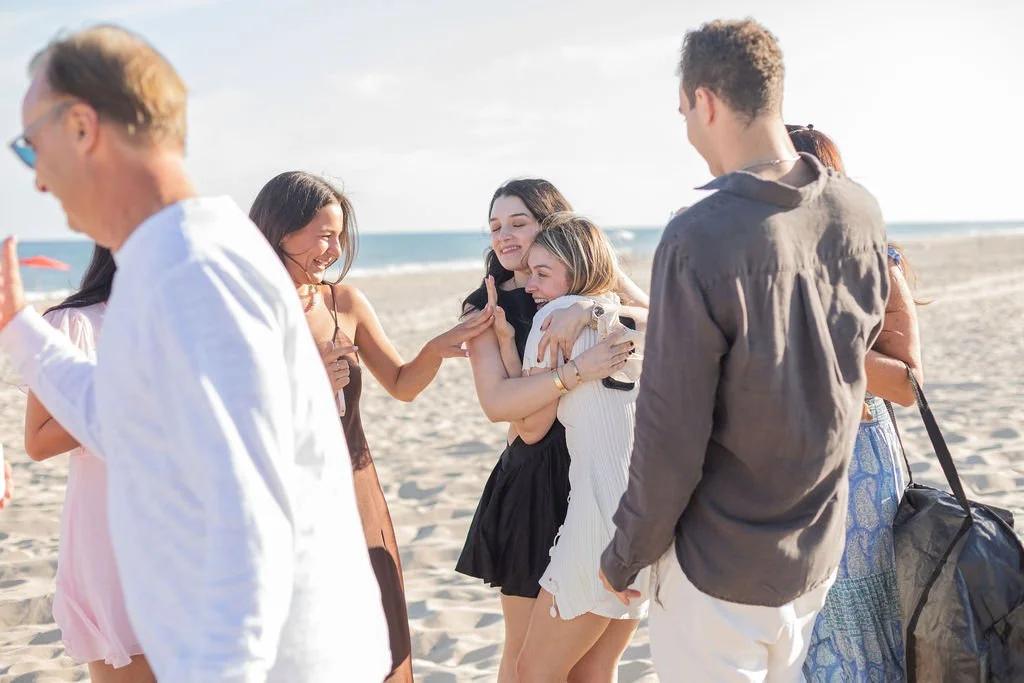 Group of friends hugging and laughing on a beach, with the ocean and sky in the background.