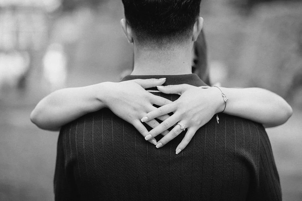 A woman hugging a man from behind with her arms wrapped around his shoulders, focusing on their embrace from the back, in black and white.