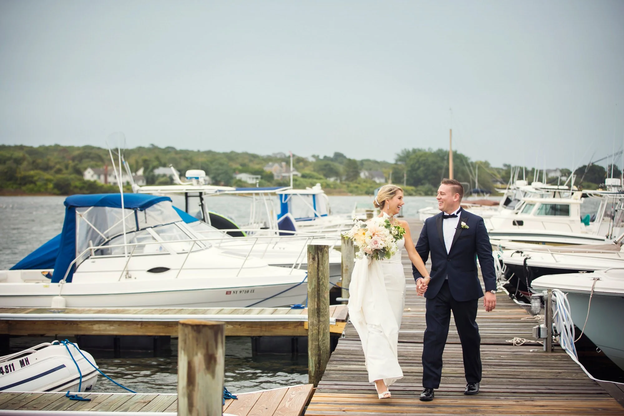 A newlywed couple walking hand in hand on a dock near a marina with boats, smiling and looking at each other, the bride holding a large bouquet of flowers, both dressed in wedding attire.